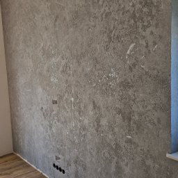 GOLD FINISH Filip Mróz - Szara ściana z efektem dekoracyjnym w Zielonej Górze, widoczne gniazdka elektryczne i drewnopodobna podłoga.