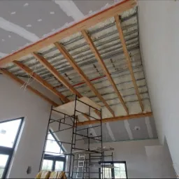Skośny sufit z widoczną konstrukcją drewnianą, izolacją piankową i częściowo zamontowaną płytą karton gips, widok z dołu na rusztowaniu.