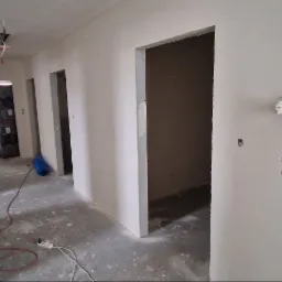 Korytarz w trakcie prac wykończeniowych, ściany przygotowane pod malowanie z widocznymi otworami na gniazdka, podłoga surowa, widoczne przewody i narzędzia.