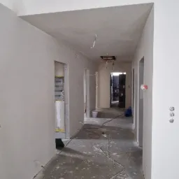 Korytarz w trakcie prac wykończeniowych, ściany i sufit pokryte świeżą warstwą gładzi, widoczne przewody elektryczne i pozostałości po pracach na podłodze, otwór w suficie bez oprawy oświetleniowej.