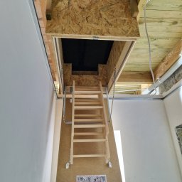 GOLD FINISH Filip Mróz - Otwór strychowy z drewnianą składaną drabinką w suficie, widoczne elementy konstrukcyjne dachu i instalacje.