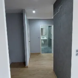 Wnętrze korytarza z panelami podłogowymi w odcieniach brązu, szarą ścianą z fakturą i białymi listwami przypodłogowymi, z widokiem na łazienkę z umywalką i okrągłym lustrem.