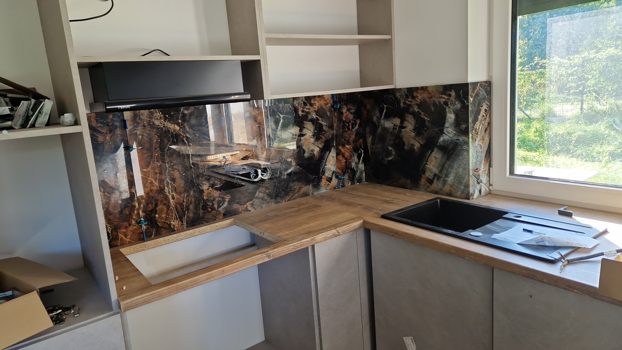 Montaż blatu kuchennego z czarnym zlewozmywakiem i panelu ściennego imitującego marmur, widoczne uchwyty mocujące panel.