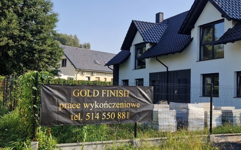 Nowy dom z czarnym dachem i białymi ścianami, widoczny baner reklamowy z napisem 'GOLD FINISH prace wykończeniowe tel. 514 550 881', na pierwszym planie stos materiałów budowlanych.