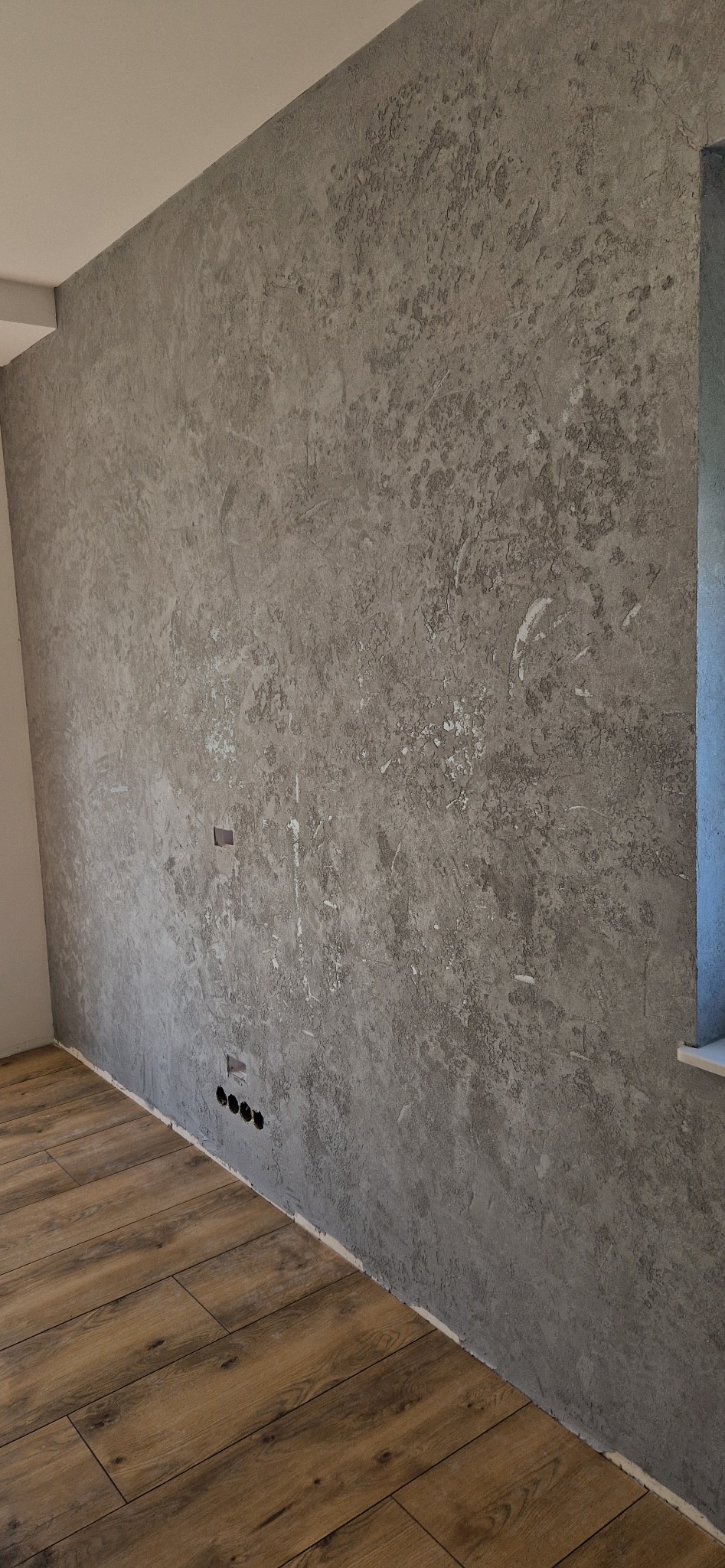 Szara ściana z efektem dekoracyjnym w Zielonej Górze, widoczne gniazdka elektryczne i drewnopodobna podłoga.