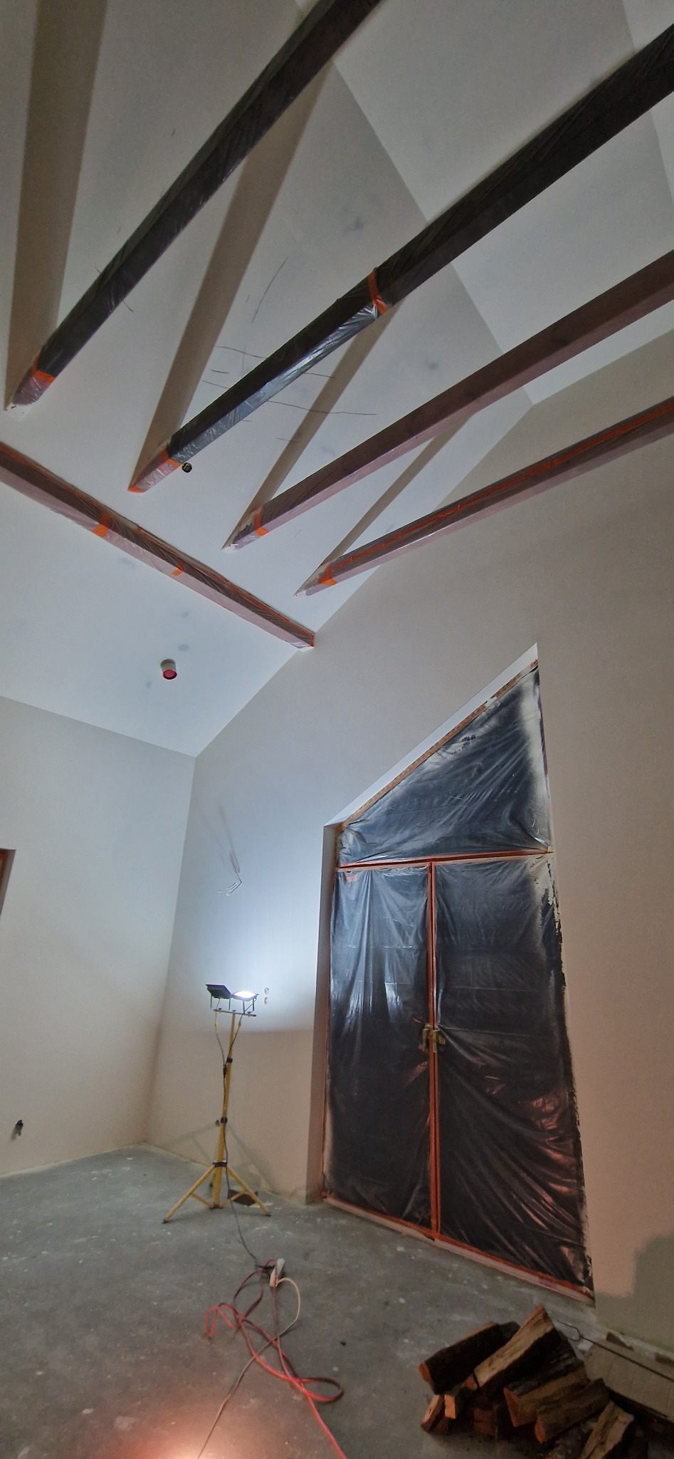 Wnętrze w trakcie prac: drewniane belki stropowe zabezpieczone folią, okno zakryte czarną folią ochronną, halogen na statywie oświetla pomieszczenie, na podłodze leżą kable i drewno.