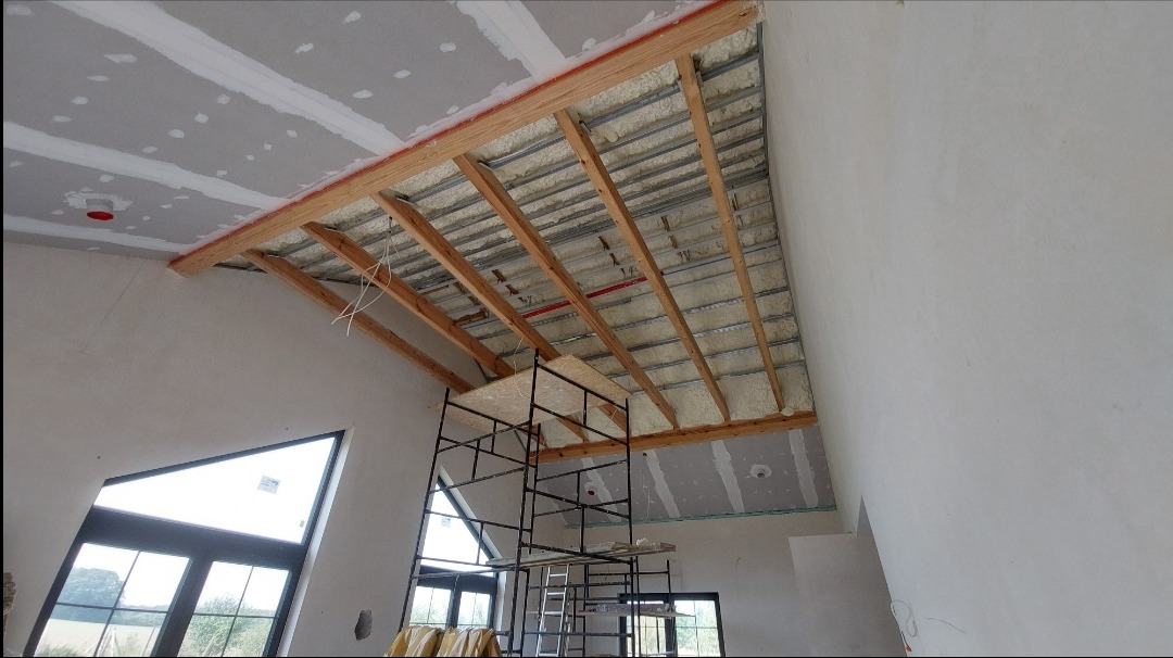 Skośny sufit z widoczną konstrukcją drewnianą, izolacją piankową i częściowo zamontowaną płytą karton gips, widok z dołu na rusztowaniu.