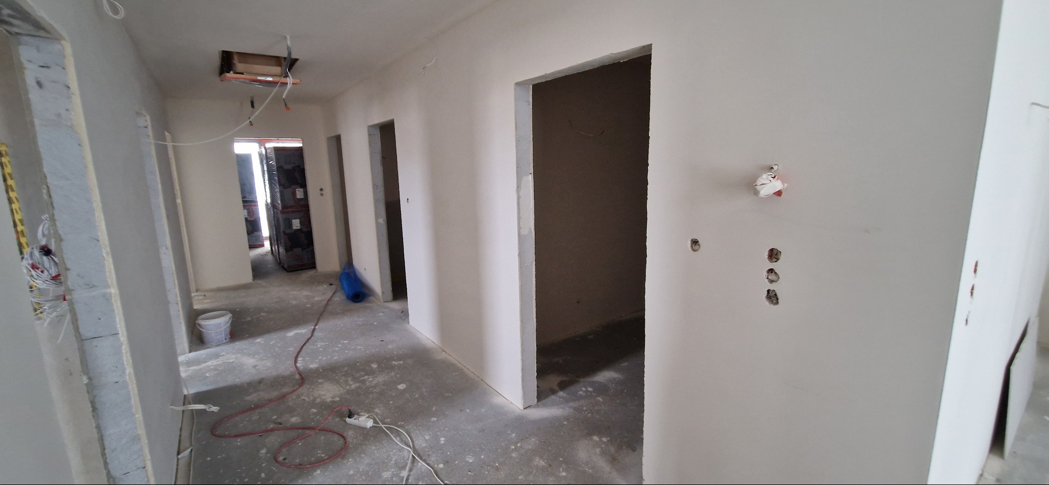 Korytarz w trakcie prac wykończeniowych, ściany przygotowane pod malowanie z widocznymi otworami na gniazdka, podłoga surowa, widoczne przewody i narzędzia.