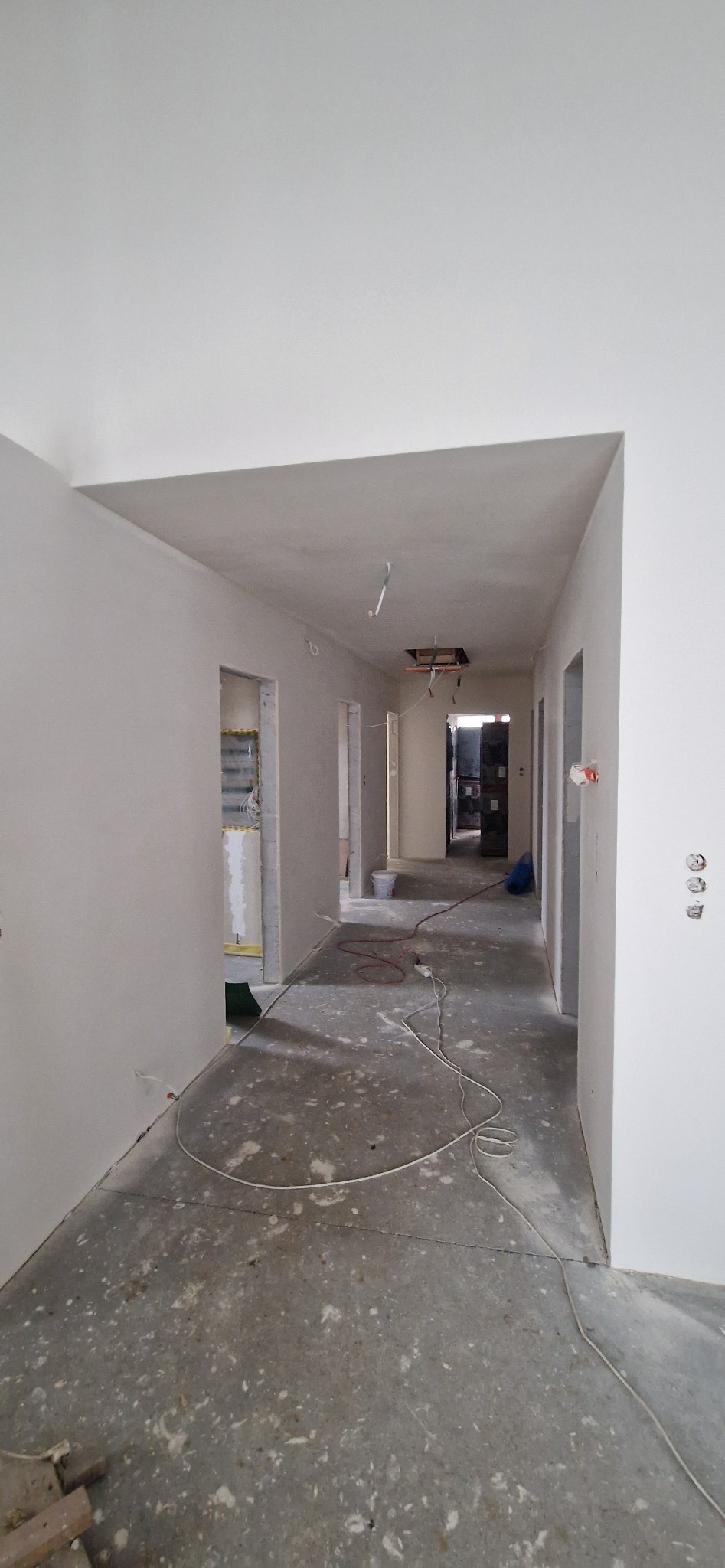 Korytarz w trakcie prac wykończeniowych, ściany i sufit pokryte świeżą warstwą gładzi, widoczne przewody elektryczne i pozostałości po pracach na podłodze, otwór w suficie bez oprawy oświetleniowej.