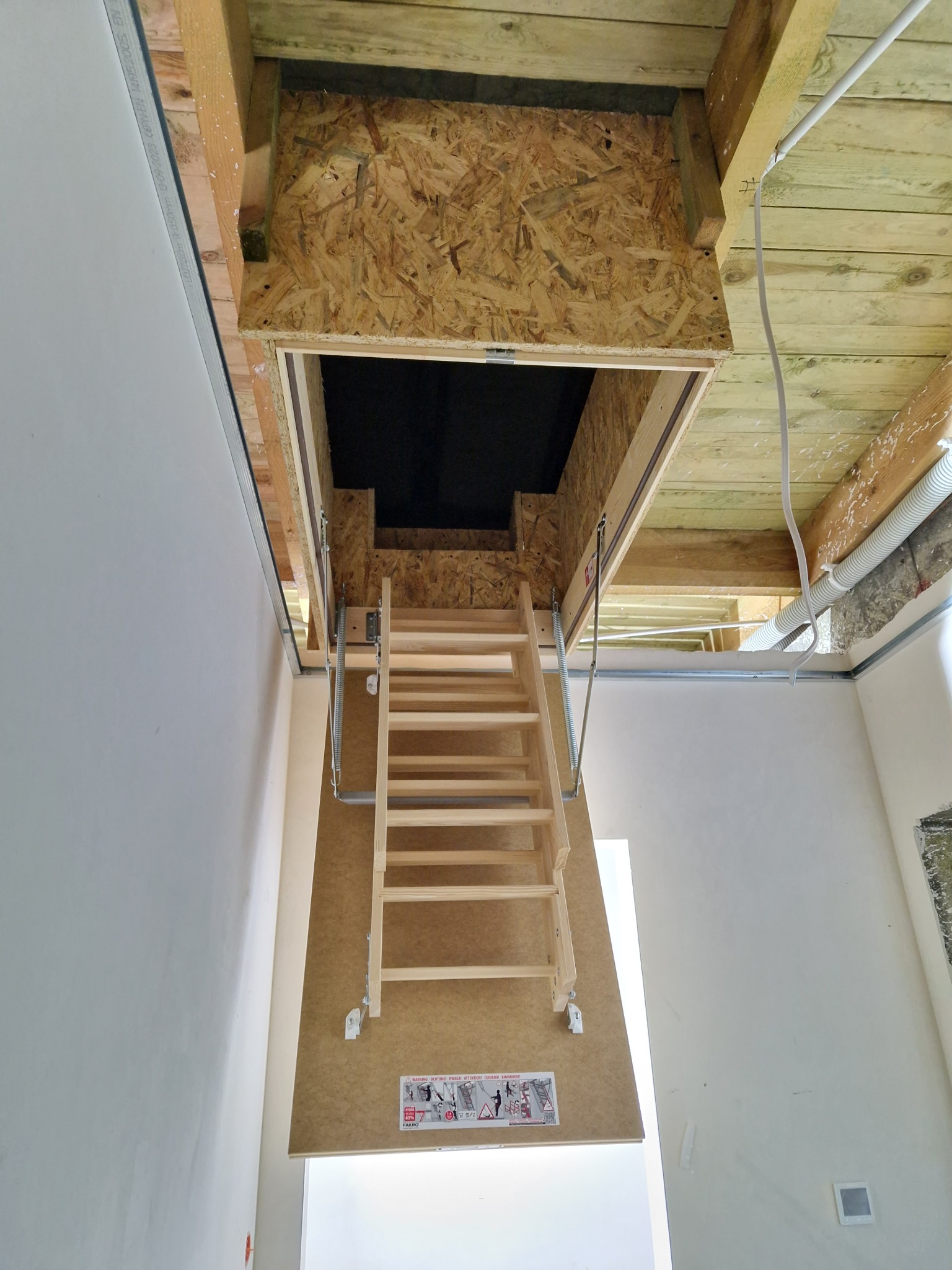 Otwór strychowy z drewnianą składaną drabinką w suficie, widoczne elementy konstrukcyjne dachu i instalacje.