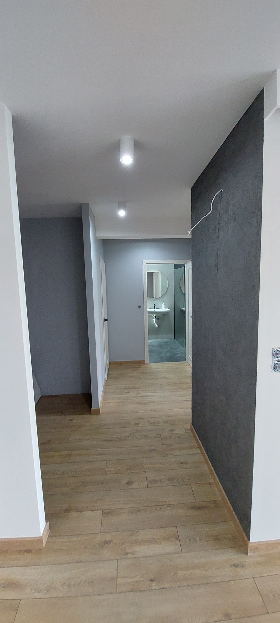 Wnętrze korytarza z panelami podłogowymi w odcieniach brązu, szarą ścianą z fakturą i białymi listwami przypodłogowymi, z widokiem na łazienkę z umywalką i okrągłym lustrem.