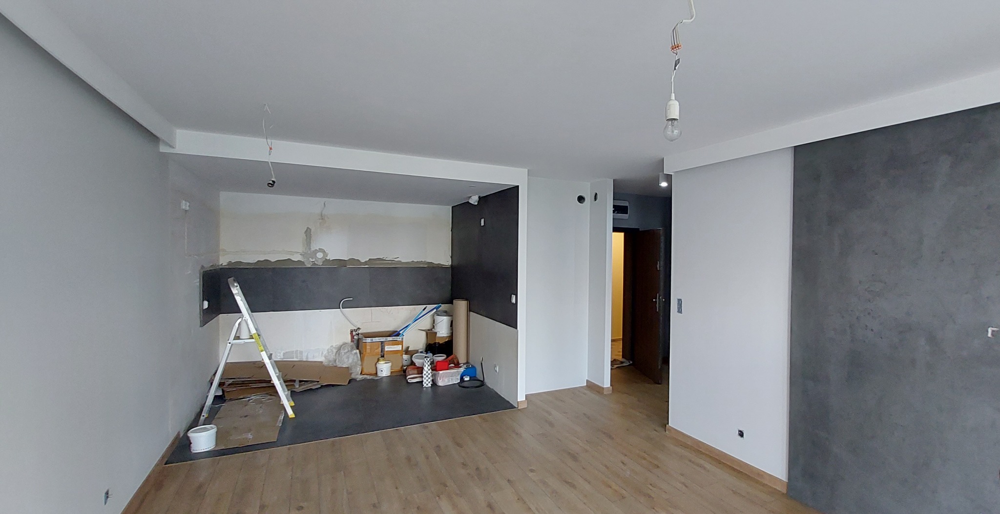 Wnętrze w trakcie remontu: drewniana podłoga, drabina oparta o ścianę, przygotowane materiały i narzędzia, fragment ściany z szarą strukturą dekoracyjną.
