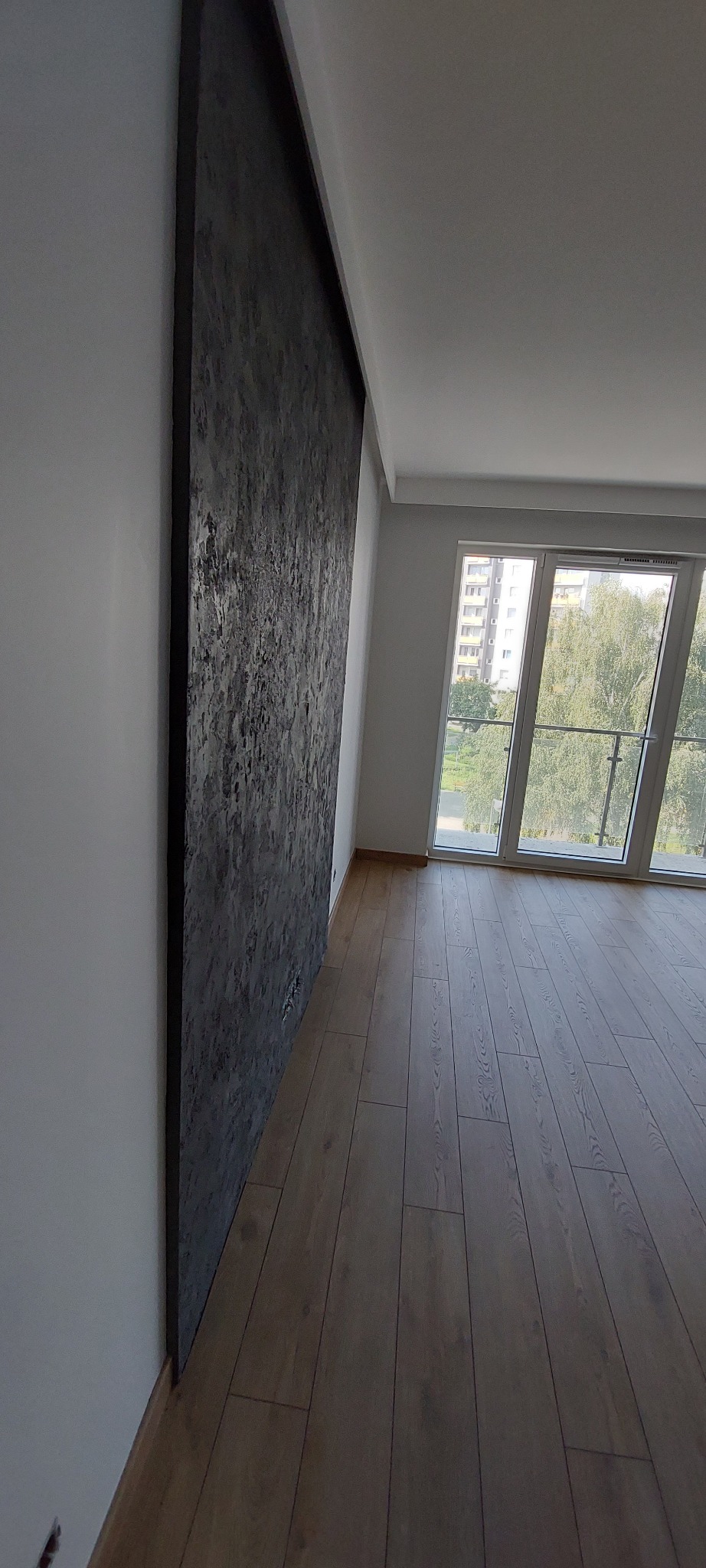 Ściana z dekoracyjną, ciemną strukturą w nowoczesnym wnętrzu z drewnianą podłogą i dużym oknem balkonowym.