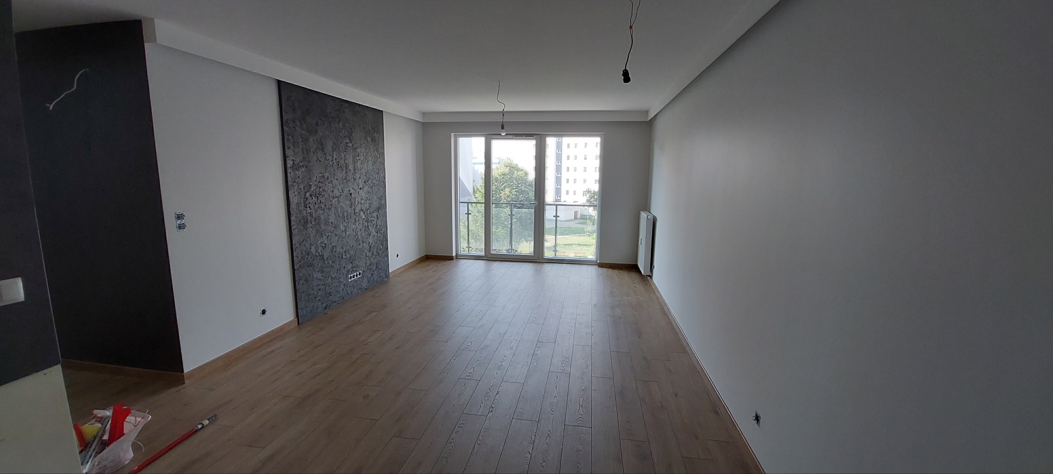 Pomalowane na szaro wnętrze mieszkania z panelami podłogowymi, ozdobną ścianą z efektem betonu i widocznymi puszkami elektrycznymi przed montażem osprzętu.