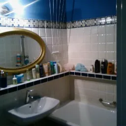 Umywalka i wanna w łazience z białymi płytkami i niebieskim sufitem, lustro w złotej ramie odbija kabinę prysznicową, a na półce stoją kosmetyki.