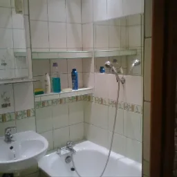 Wyremontowana łazienka z białą wanną, umywalką i prysznicem. Ściany wyłożone białymi płytkami z dekoracyjnym wzorem. Widoczne półki z kosmetykami i fragment ściany wykończonej kamieniem dekoracyjnym.