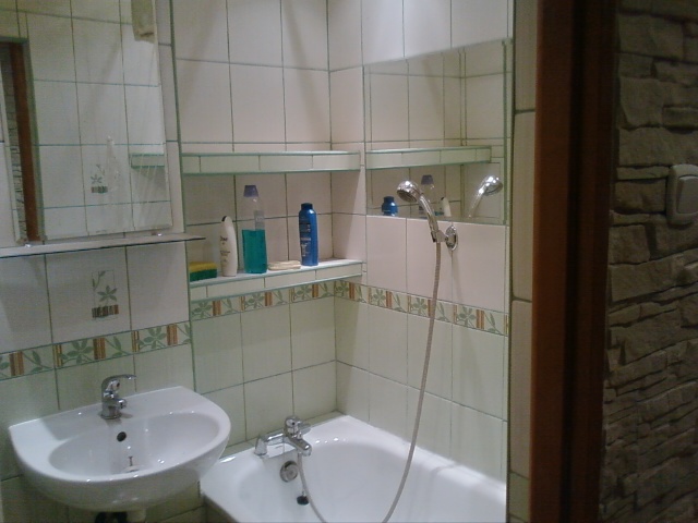 Wyremontowana łazienka z białą wanną, umywalką i prysznicem. Ściany wyłożone białymi płytkami z dekoracyjnym wzorem. Widoczne półki z kosmetykami i fragment ściany wykończonej kamieniem dekoracyjnym.