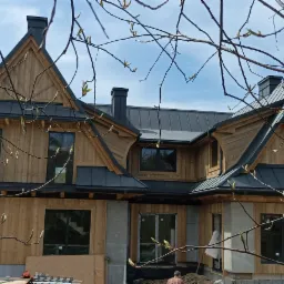 Dom w stylu zakopiańskim z drewnianą elewacją i szarą blachą na dachu, widok z dołu przez gałęzie drzew z pąkami, w tle błękitne niebo.