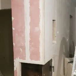 Niedokończona obudowa kominka z wkładem, widoczne płyty gipsowo-kartonowe, szpachlowanie i czarne wstawki wentylacyjne, na podłodze resztki materiałów budowlanych, drabina oparta o ścianę.
