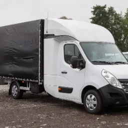 Biały samochód dostawczy z czarną plandeką na pakę, zaparkowany na żwirowym parkingu, widok z boku.