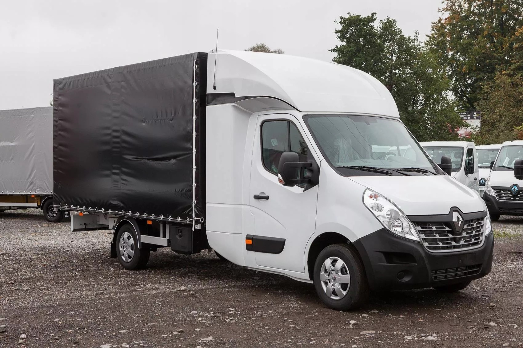 Biały samochód dostawczy z czarną plandeką na pakę, zaparkowany na żwirowym parkingu, widok z boku.