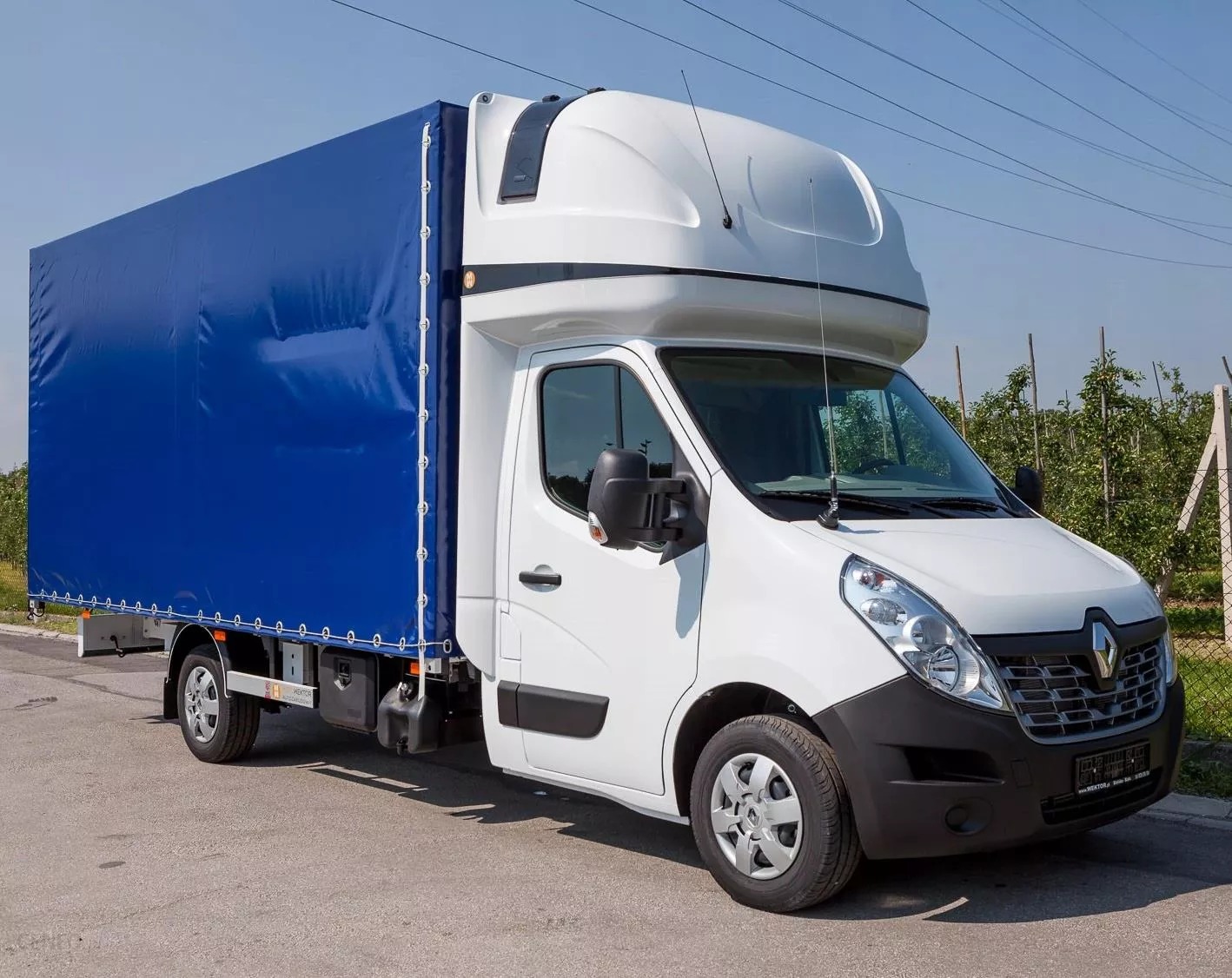 Biały samochód dostawczy Renault z niebieską plandeką na parkingu, widok z przodu i boku, antena na dachu, w tle drzewa owocowe.