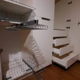 Wnętrze szafy wnękowej z wysuwanymi koszami drucianymi i półkami, widoczne elementy montażowe na ścianie, drewniana podłoga.