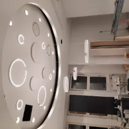 Nowoczesny sufit podwieszany z okrągłymi, podświetlanymi elementami i wiszącymi lampami pierścieniowymi, widoczne okna i elementy konstrukcyjne w trakcie montażu.