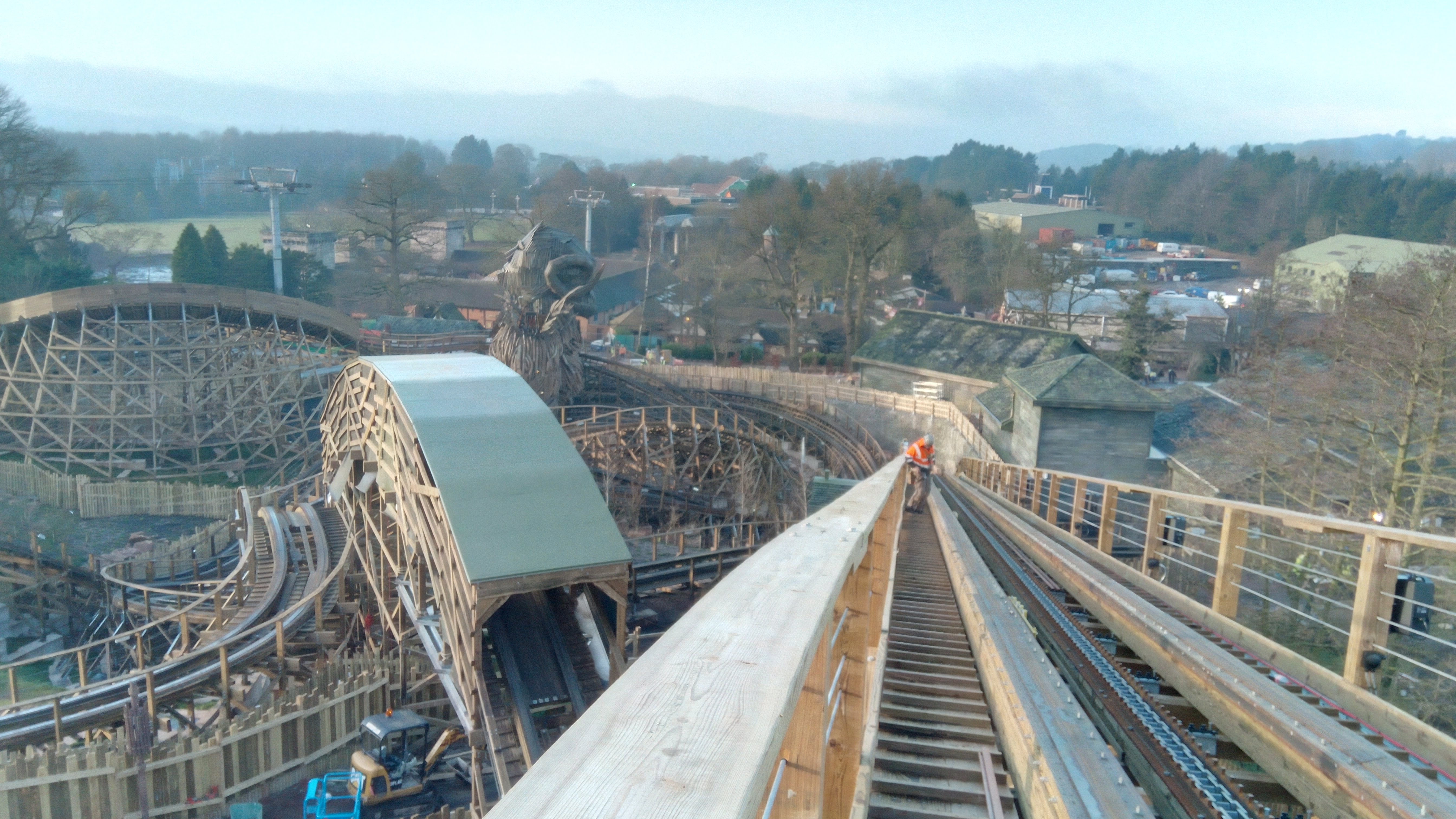 Konstrukcja drewnianego rollercoastera w parku rozrywki, z pracownikiem w pomarańczowej kamizelce na torze, w tle widoczna głowa zwierzęcia i zabudowania.