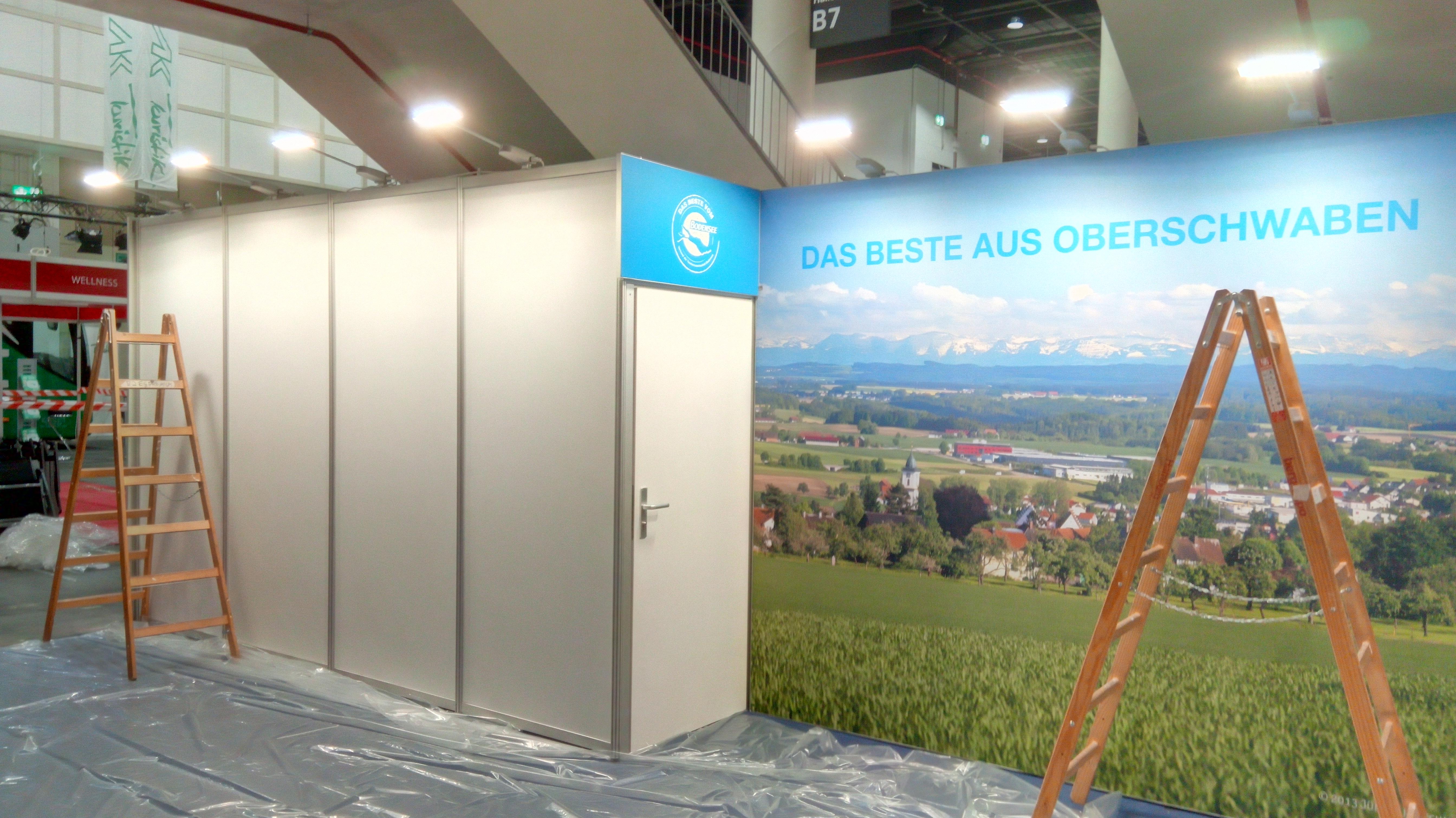 Przygotowanie stoiska targowego w Nowym Stawie: drabina oparta o ściankę działową z drzwiami i fototapetą przedstawiającą krajobraz wiejski z górami w tle, podłoga zabezpieczona folią.