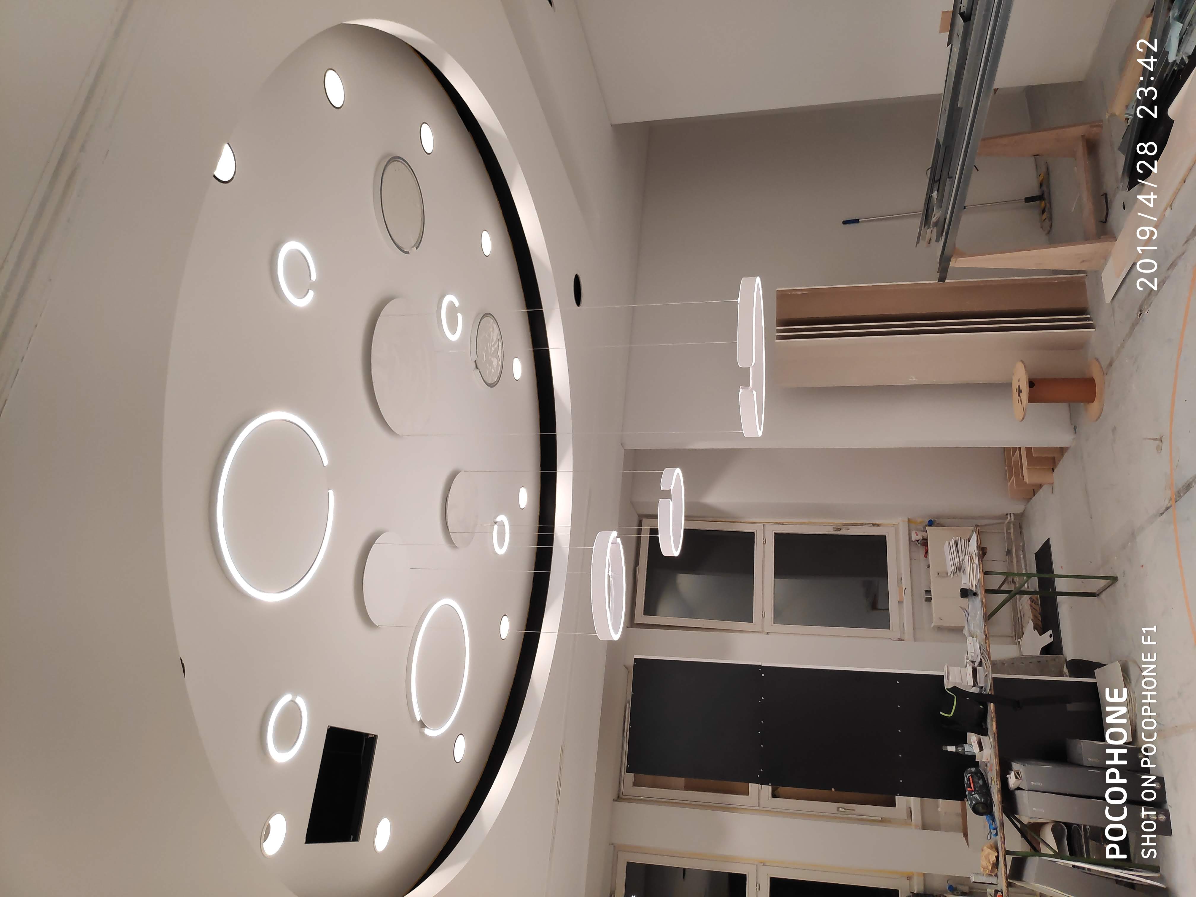 Nowoczesny sufit podwieszany z okrągłymi, podświetlanymi elementami i wiszącymi lampami pierścieniowymi, widoczne okna i elementy konstrukcyjne w trakcie montażu.