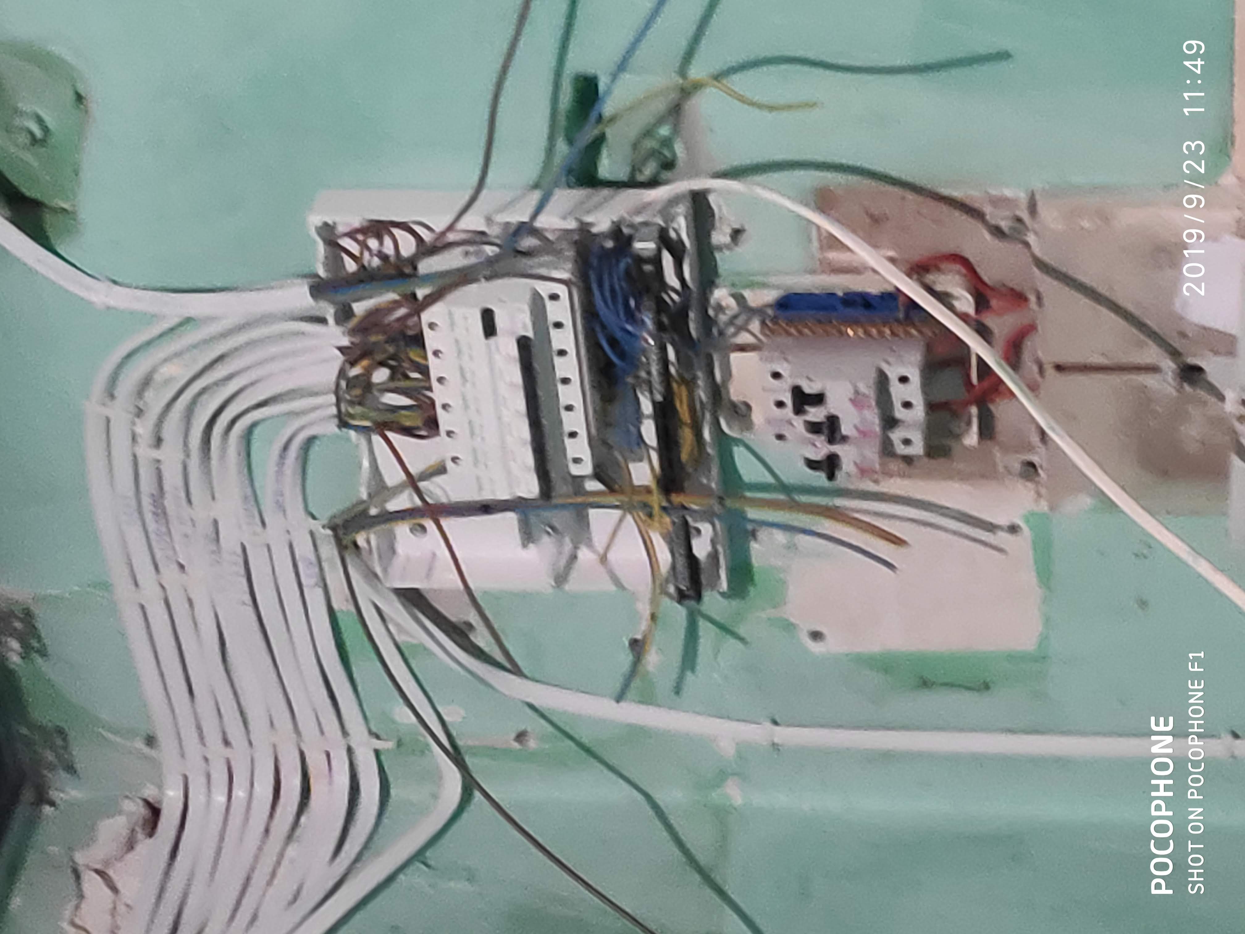 Otwarta skrzynka elektryczna z licznymi przewodami w ścianie pomalowanej na zielono, widoczne elementy złączek i bezpieczników.