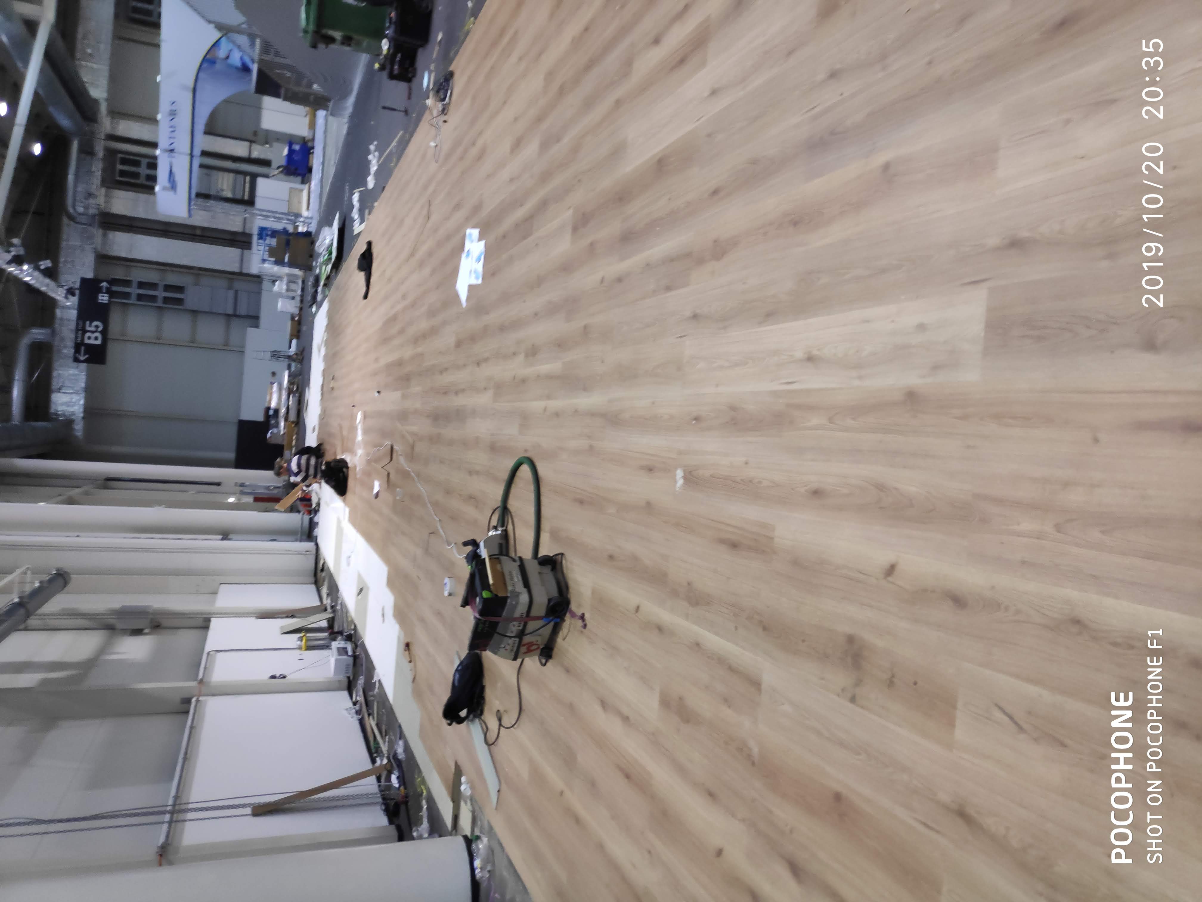 Maszyna do czyszczenia posadzek przemysłowych na jasnej, drewnopodobnej podłodze w hali, widoczny fragment białej ściany i konstrukcji sufitu.