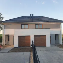 Nowoczesny, dwukondygnacyjny dom bliźniak z szarą elewacją i brązowymi bramami garażowymi, widok z zewnątrz, ułożona kostka brukowa i przygotowane do ułożenia obrzeża.