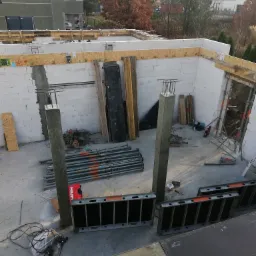 Wysoki kąt widzenia na budowę domu jednorodzinnego w stanie surowym otwartym, widoczne ściany z bloczków, słupy żelbetowe z wystającymi prętami zbrojeniowymi, podpory stalowe i szalunki.