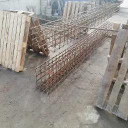 Dwie prostokątne, przestrzenne konstrukcje z prętów zbrojeniowych, ułożone równolegle na ziemi, wsparte na drewnianych paletach, z widocznym ciężarowym samochodem w tle.