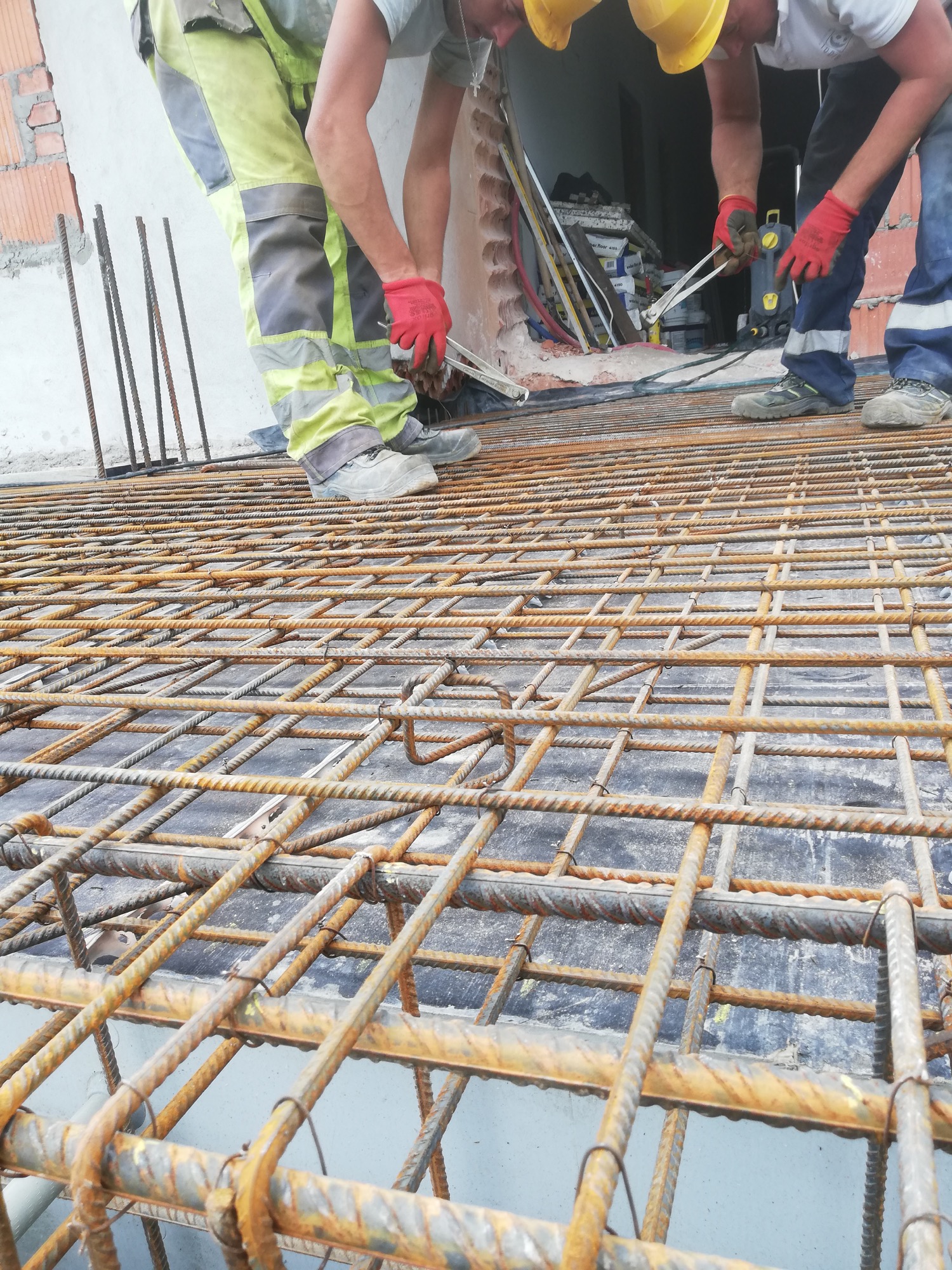 Praca na budowie: trzech robotników w kaskach i rękawicach łączy stalowe pręty zbrojeniowe na przygotowanym szalunku, widoczne narzędzia i materiały budowlane w tle.