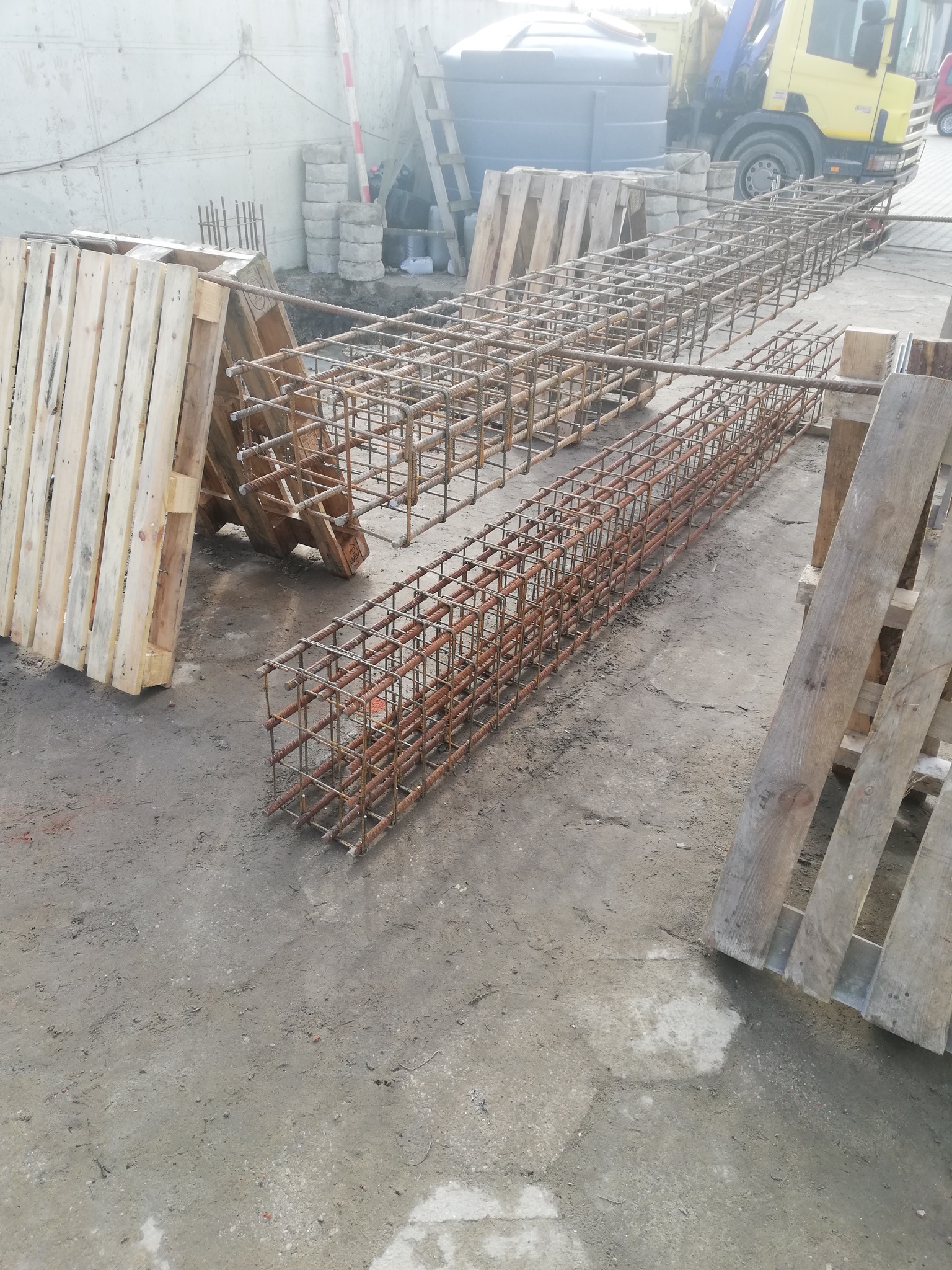 Dwie prostokątne, przestrzenne konstrukcje z prętów zbrojeniowych, ułożone równolegle na ziemi, wsparte na drewnianych paletach, z widocznym ciężarowym samochodem w tle.