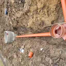 Wykonany wykop z ułożonymi pomarańczowymi rurami kanalizacyjnymi, studzienka betonowa z czerwonymi szmatami w środku, szpadel w ziemi, widok z g&oacute;ry.