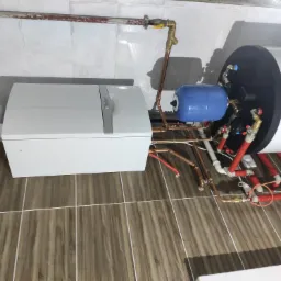 Biały piec grzewczy Vaillant z podłączonymi rurami miedzianymi i zbiornikiem wyrównawczym, zamontowany na brązowych płytkach podłogowych w jasnym pomieszczeniu.