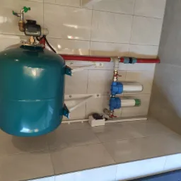 Niebiesko-zielony zbiornik hydroforowy z manometrem i zaworem, zamontowany na ścianie z kafelków, widoczne filtry wody, rury PCV i przewody elektryczne.