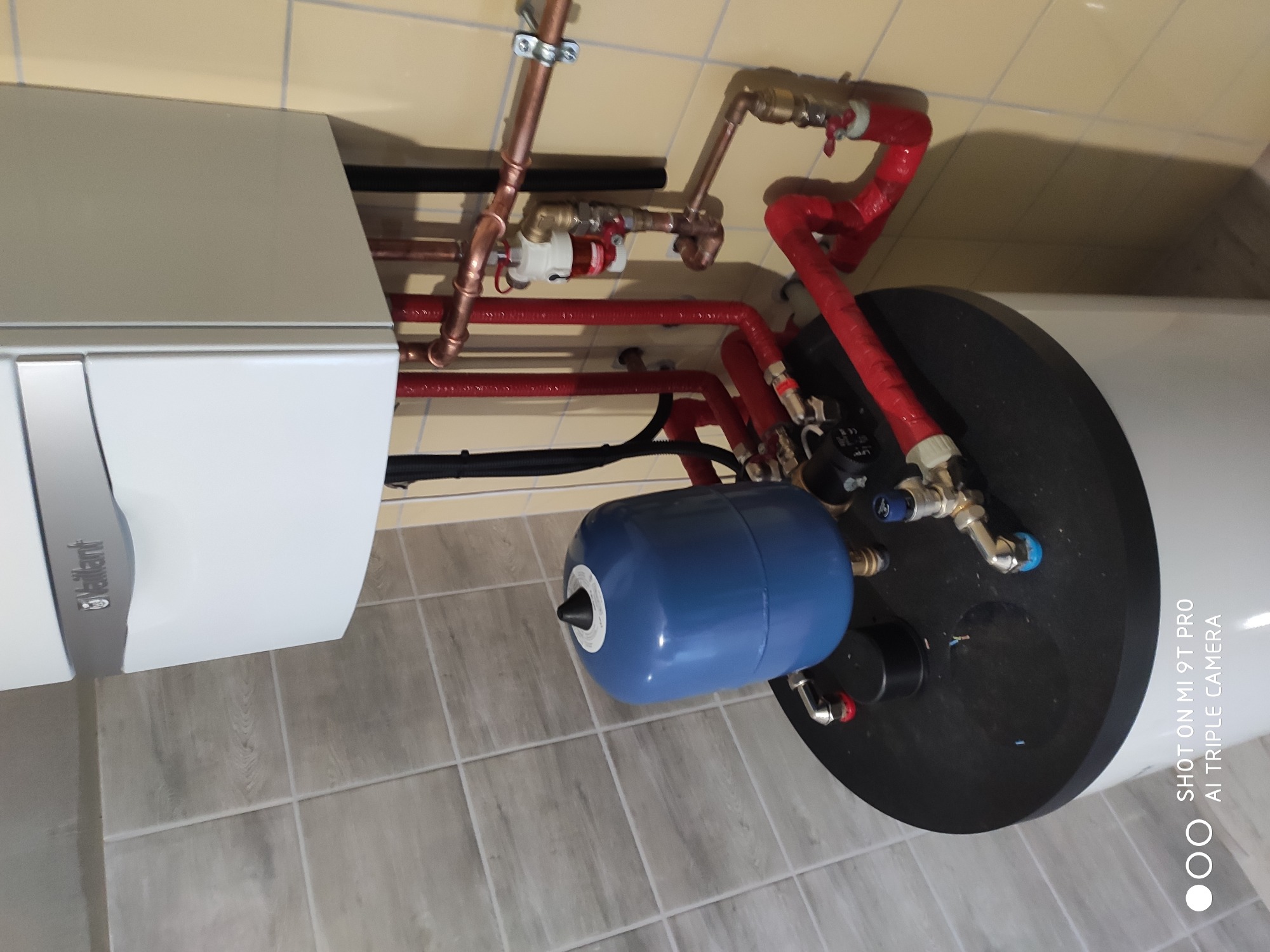 Biały piec grzewczy Vaillant z niebieskim naczyniem przeponowym i miedzianymi rurami w czerwonej izolacji, widoczne elementy przyłączeniowe.