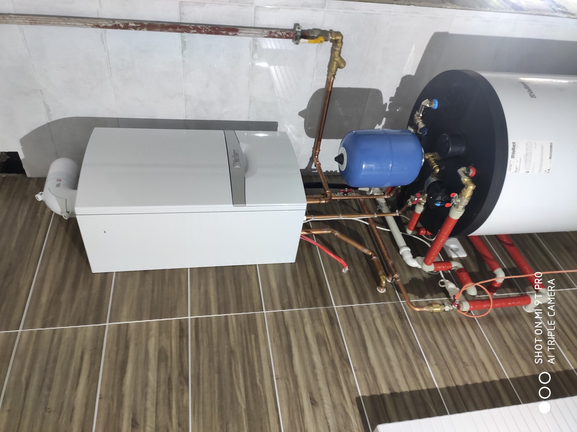 Biały piec grzewczy Vaillant z podłączonymi rurami miedzianymi i zbiornikiem wyrównawczym, zamontowany na brązowych płytkach podłogowych w jasnym pomieszczeniu.