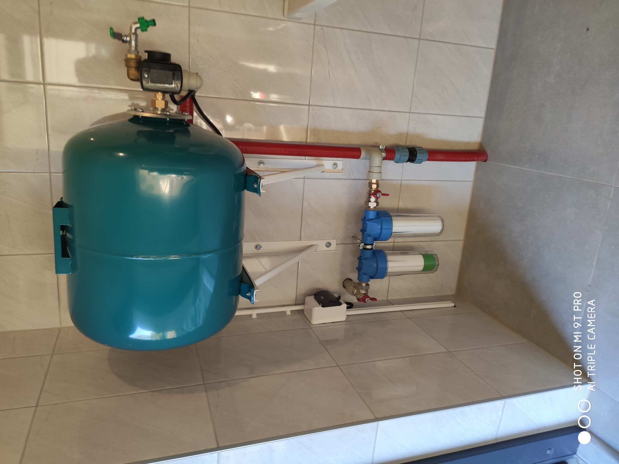 Niebiesko-zielony zbiornik hydroforowy z manometrem i zaworem, zamontowany na ścianie z kafelków, widoczne filtry wody, rury PCV i przewody elektryczne.