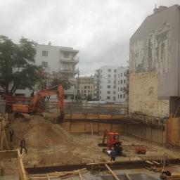 Usługi Transportowe, Wykopy, Rozbiórki, Handel - Pomarańczowa koparka pracuje przy wykopie zabezpieczonym drewnianą konstrukcją, w tle budynki mieszkalne w pochmurny dzień.