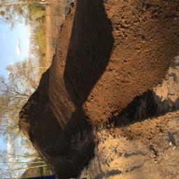 Usługi Transportowe, Wykopy, Rozbiórki, Handel - Wysoka pryzma brązowej ziemi ogrodowej, częściowo zacieniona, z widocznymi śladami opon i drzewami w tle pod błękitnym niebem.