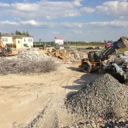 Prace rozbiórkowe z użyciem koparki i przesiewacza gruzu na placu budowy, widoczne zbrojenia i stosy gruzu.