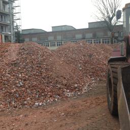 Usługi Transportowe, Wykopy, Rozbiórki, Handel - Stos gruzu ceglanego i ziemi z fragmentem żółto-czerwonej ładowarki na pierwszym planie, w tle budynek mieszkalny w trakcie budowy i budynek przemysłowy.