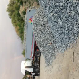 Ciężarówka z kolorowym logo wyładowuje dużą pryzmę szarego kruszywa na piaszczystym podłożu, w tle widoczna zieleń i fragment tęczy.