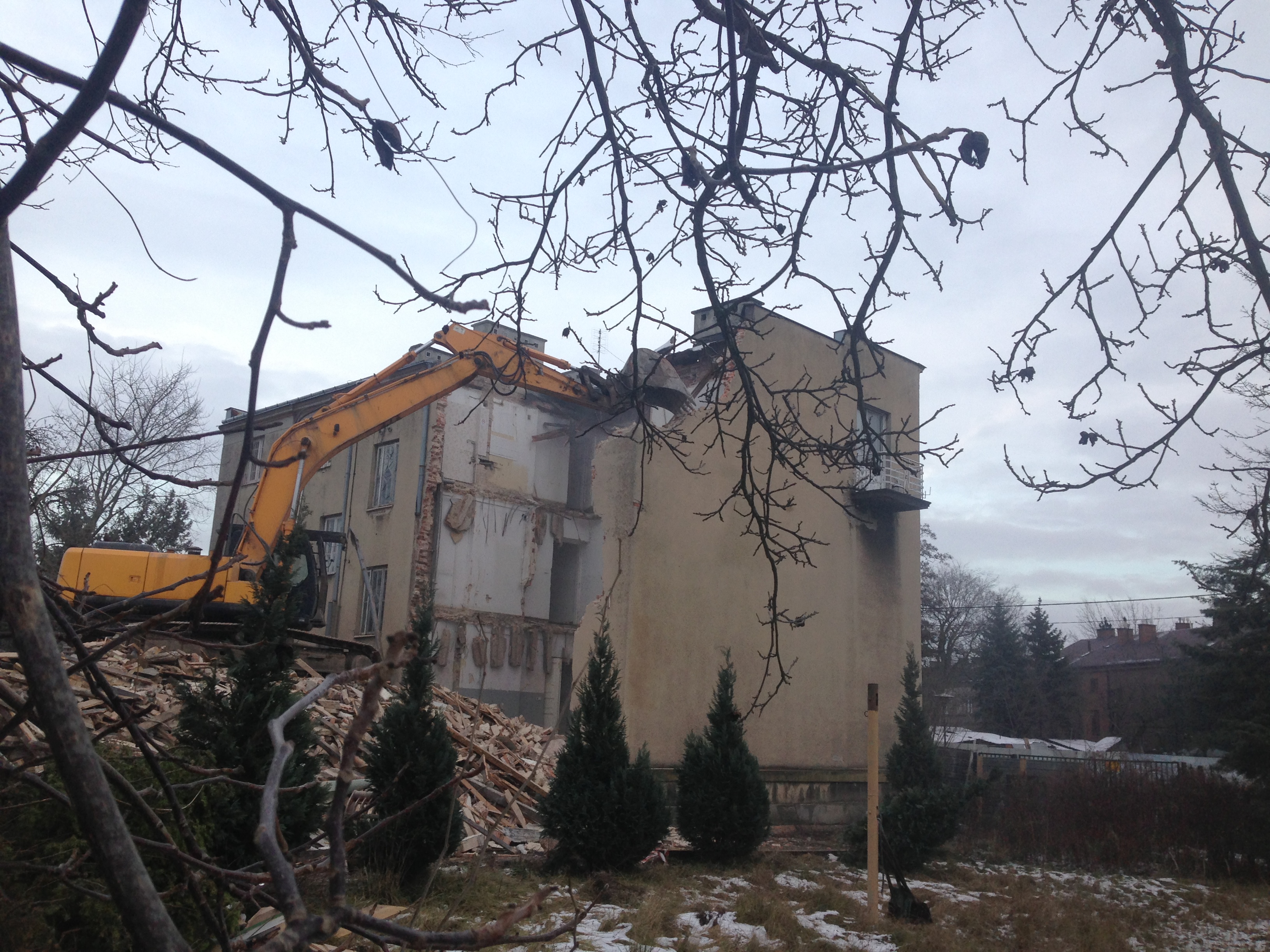Żółta koparka z wysięgnikiem w trakcie rozbiórki budynku, widoczny gruz i fragmenty konstrukcji, zima, pochmurne niebo, gałęzie na pierwszym planie.